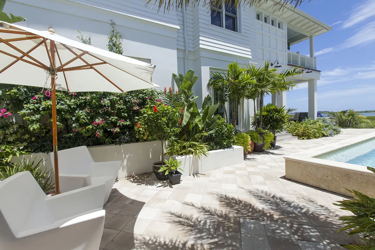 Patio with white umbrella, chairs, potted plants, stone tiles, and a swimming pool in the background.