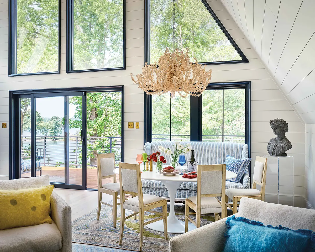 Dining room with round table, wooden chairs, chandelier, sofa, and large windows showcasing outside views