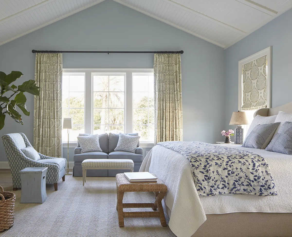 Bedroom with white quilt bed, blue chair, gray sofa, large windows, rattan stool, and light blue walls.