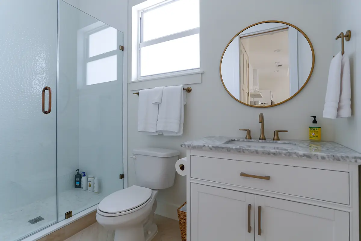 Bathroom with glass shower, white toilet, marble vanity, round mirror, and towel rack with natural light