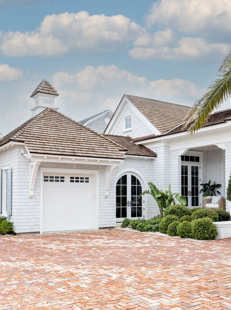 Exterior of a house with white facade, shingle roof, brick driveway, and manicured hedges.