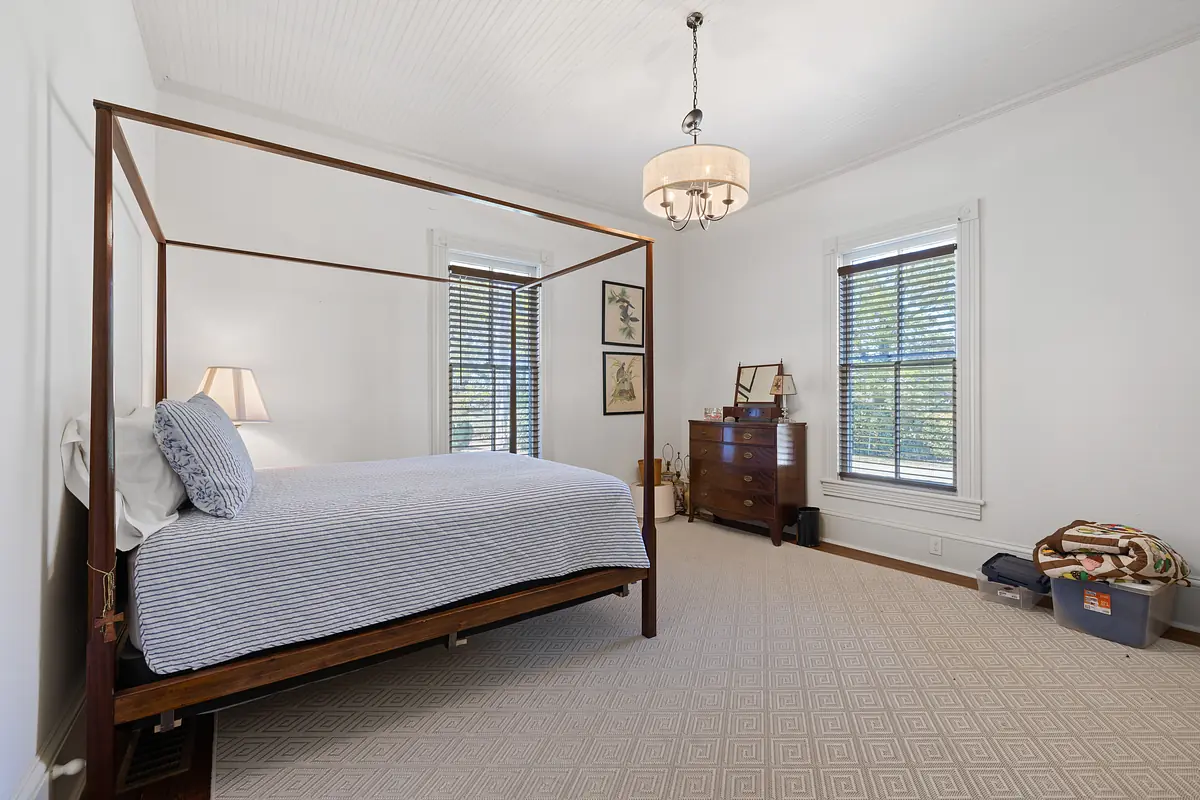 Bedroom with four-poster bed, blue striped bedding, nightstand, wooden dresser, and two windows.