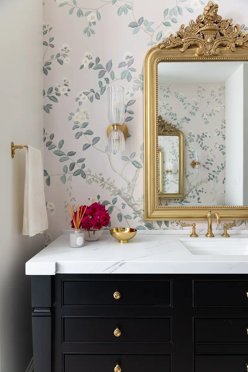 Bathroom with black vanity, gold accents, marble countertop, ornate mirror, floral wallpaper, and decorative vase