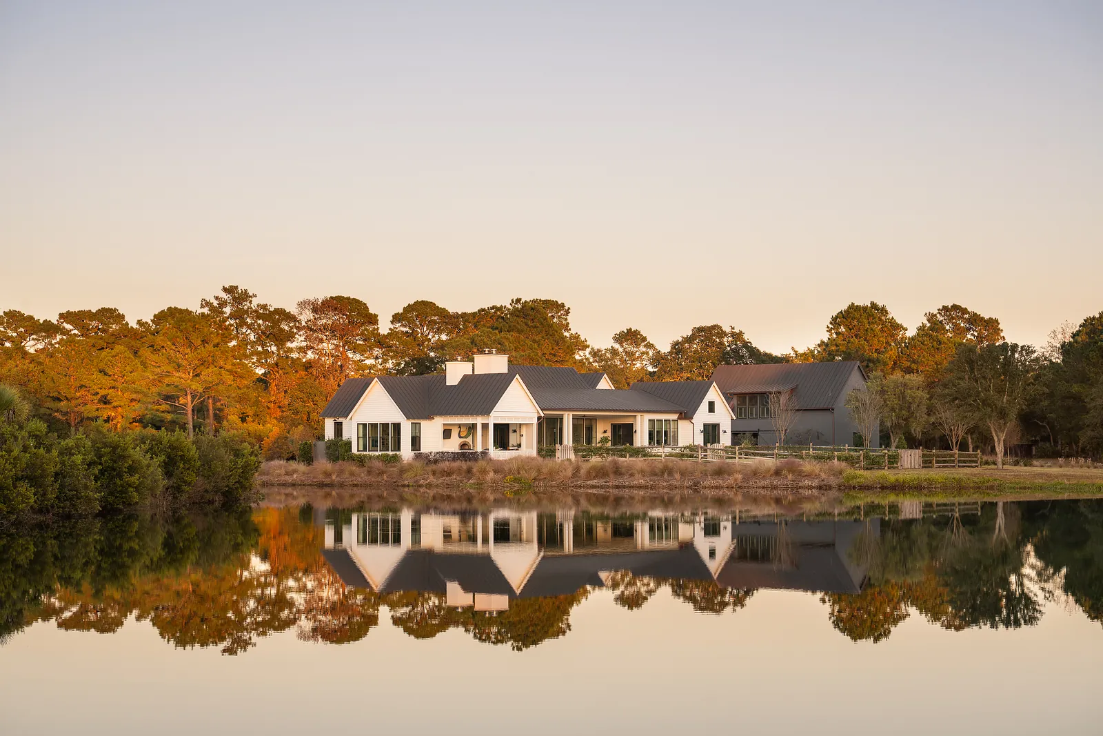 Exterior front view of a house with white facade, gray roof, large windows, and surrounded by trees and pond.