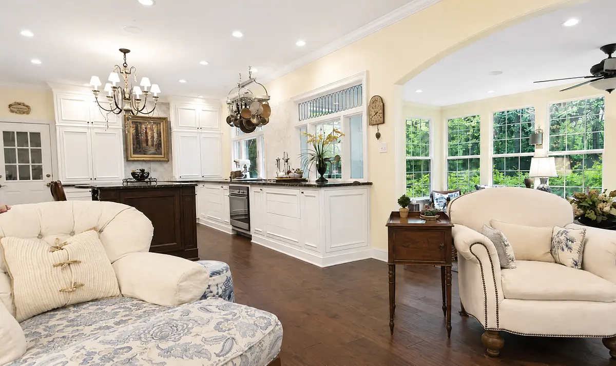 Kitchen with white cabinetry, dark countertop island, upholstered chairs, and large windows providing natural light.