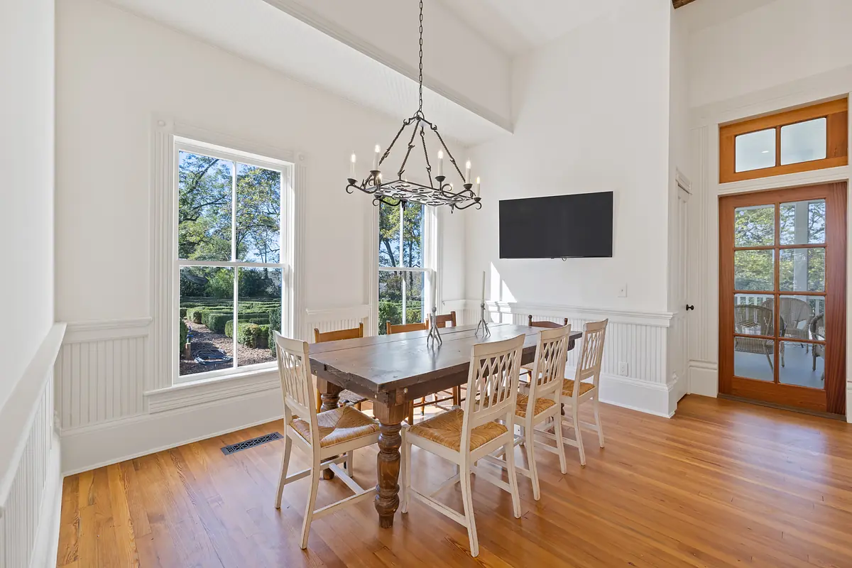 Dining room with wooden table, six chairs, chandelier, wall-mounted TV, and large windows.