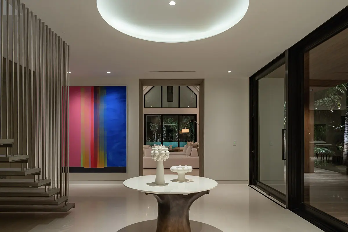 Foyer with round table, abstract painting, large windows, and staircase with metal railings.