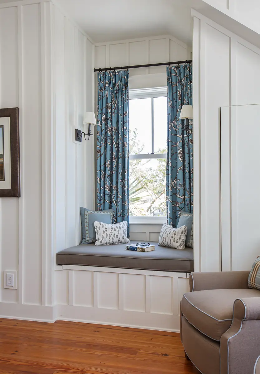 Nook with built-in window seat, blue curtains, pillows, small book, and light fixture in white paneled walls.