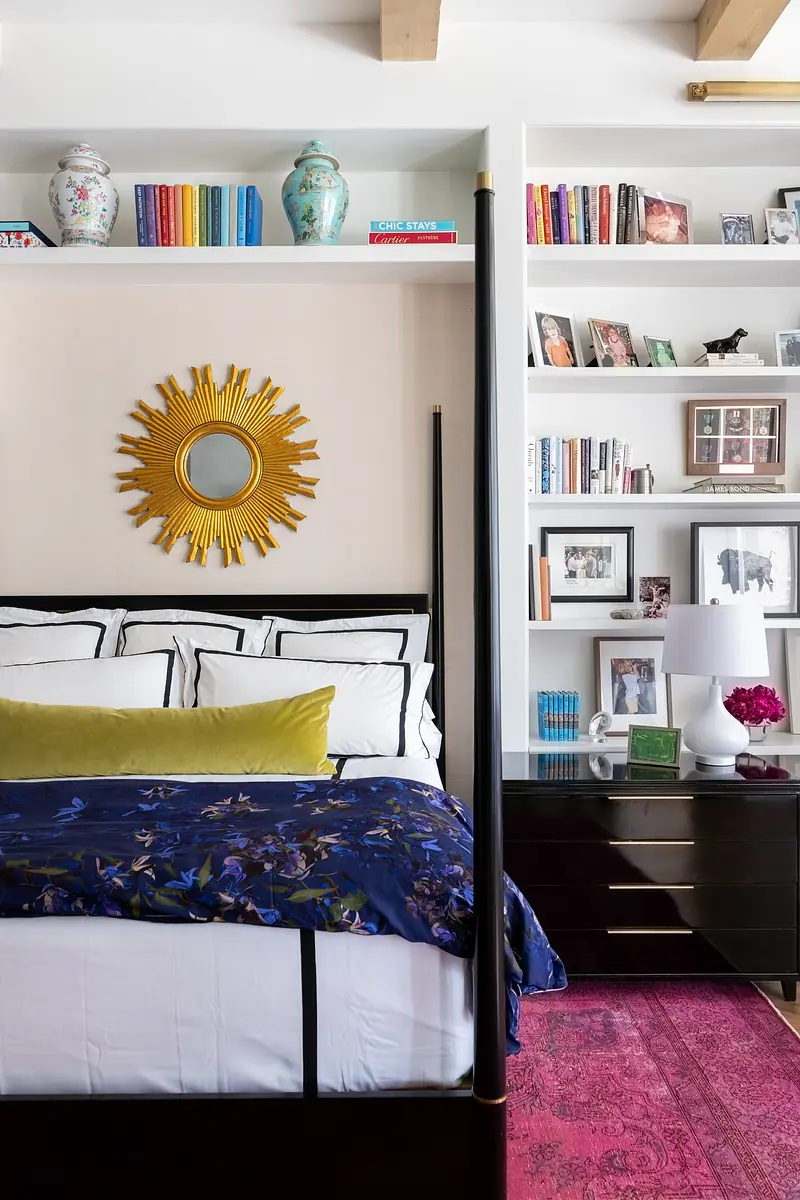 Bedroom with canopy bed, blue quilt, gold mirror, black dresser, bookshelves, and pink area rug.