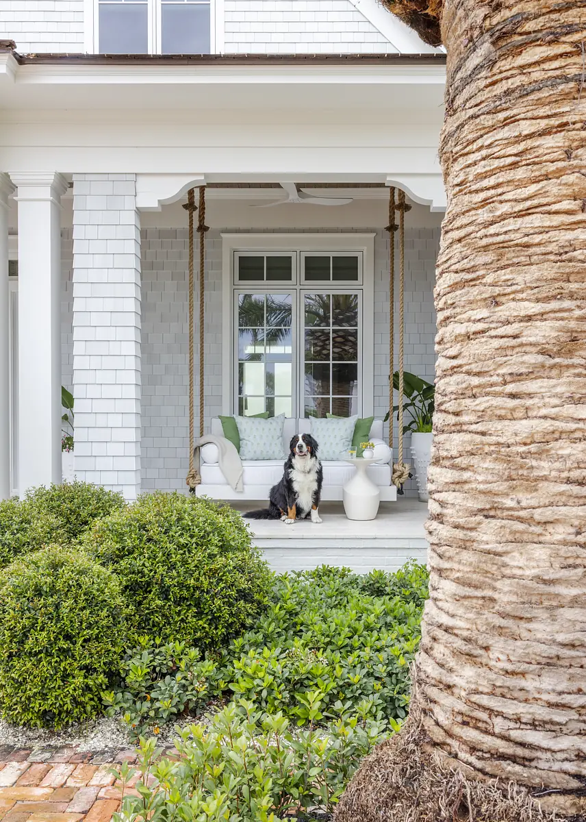 Exterior front area with porch, swing seat, plants, dog, brick flooring, and trimmed bushes.