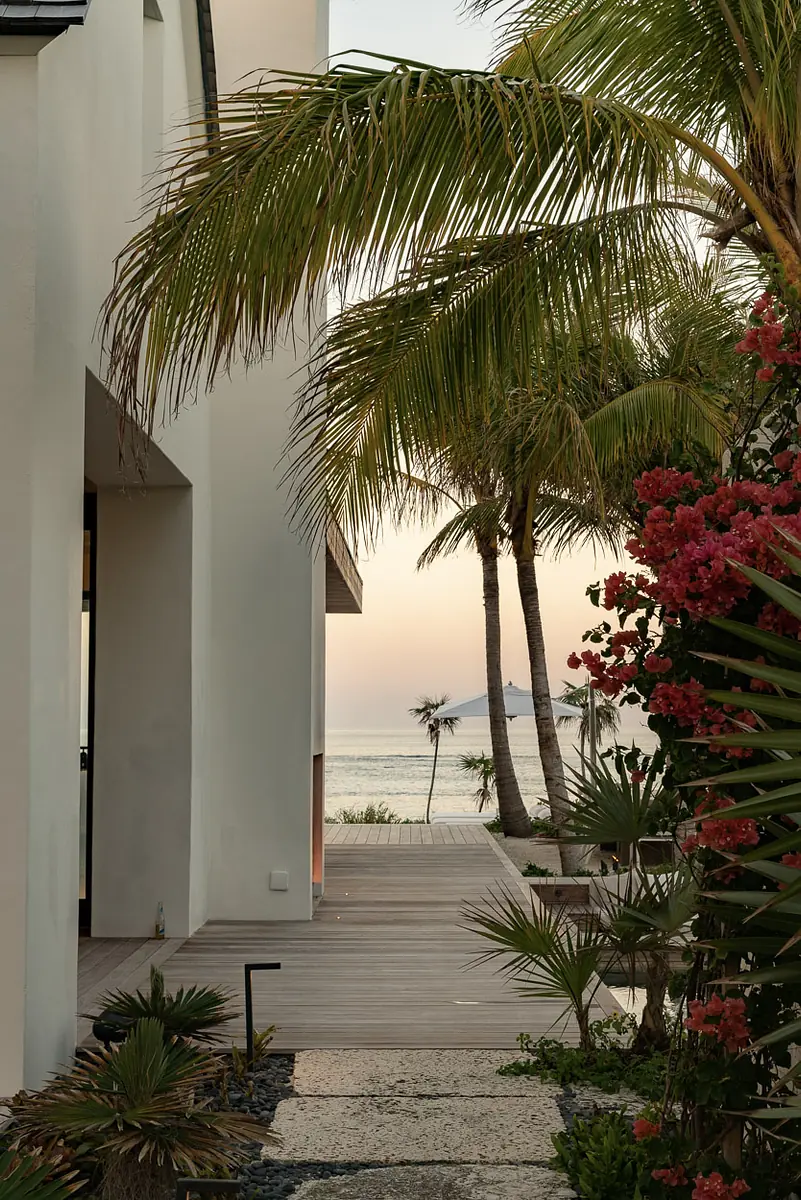 Exterior front view with wooden pathway, palm trees, and ocean in the background at sunset.
