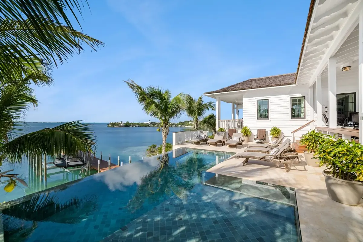 Outdoor area with pool, lounge chairs, palm trees, and house overlooking water.
