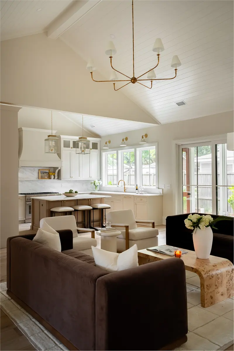 Living room with dark sofa, white armchairs, vaulted ceiling, large windows, and kitchen area in the background.
