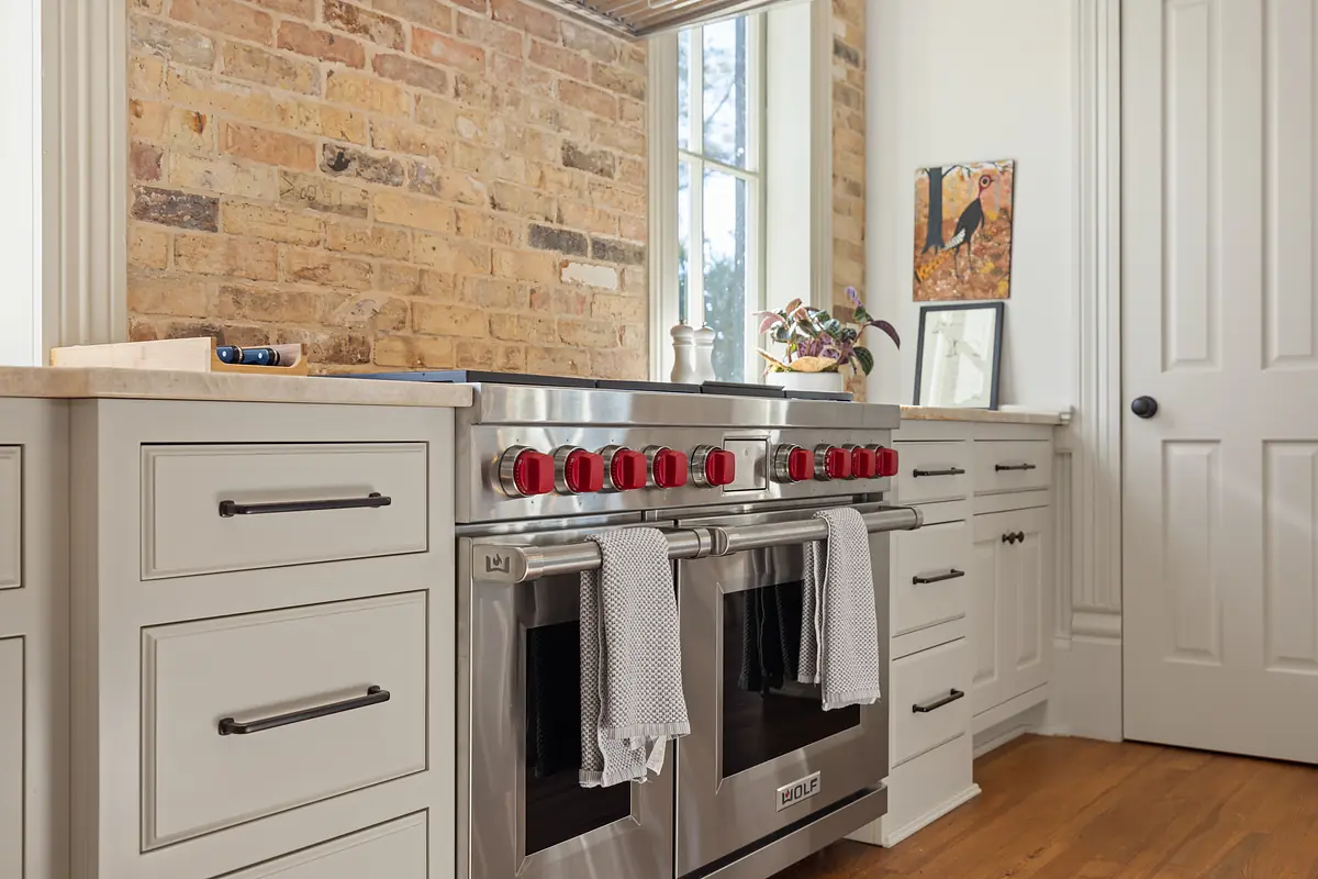 Kitchen with stainless steel range, white cabinetry, stone countertop, and exposed brick walls with large windows.