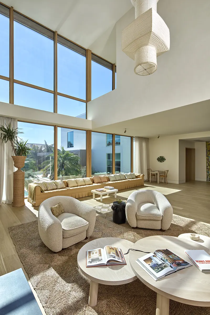 Living room with large windows, yellow sectional sofa, white armchairs, area rug, and plant.
