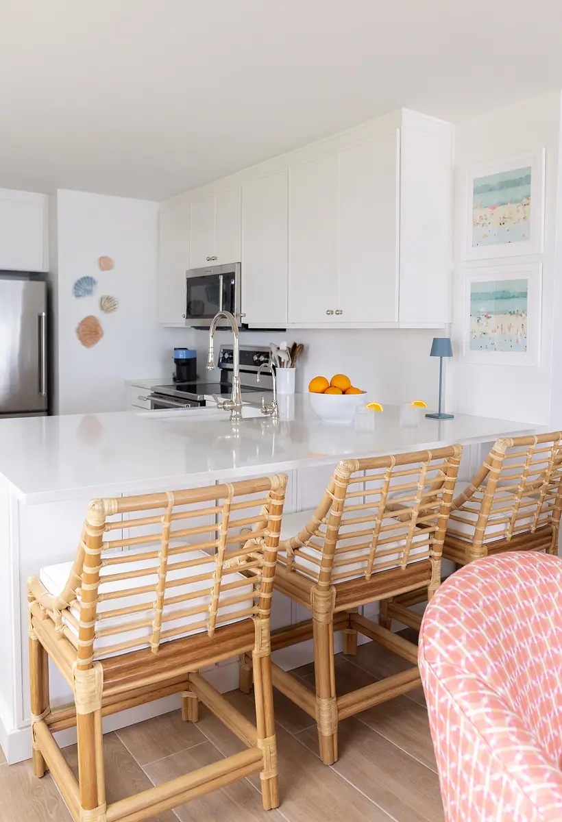 Kitchen with white cabinetry, stainless steel fridge, rattan bar stools, and a bowl of oranges on the island.