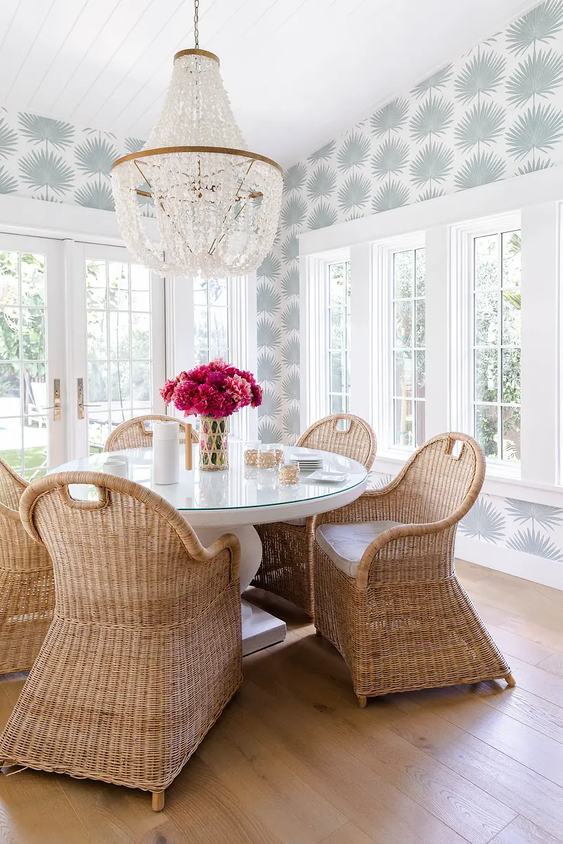 Dining room with round glass table, wicker chairs, palm leaf wallpaper, chandelier, and pink flower centerpiece.