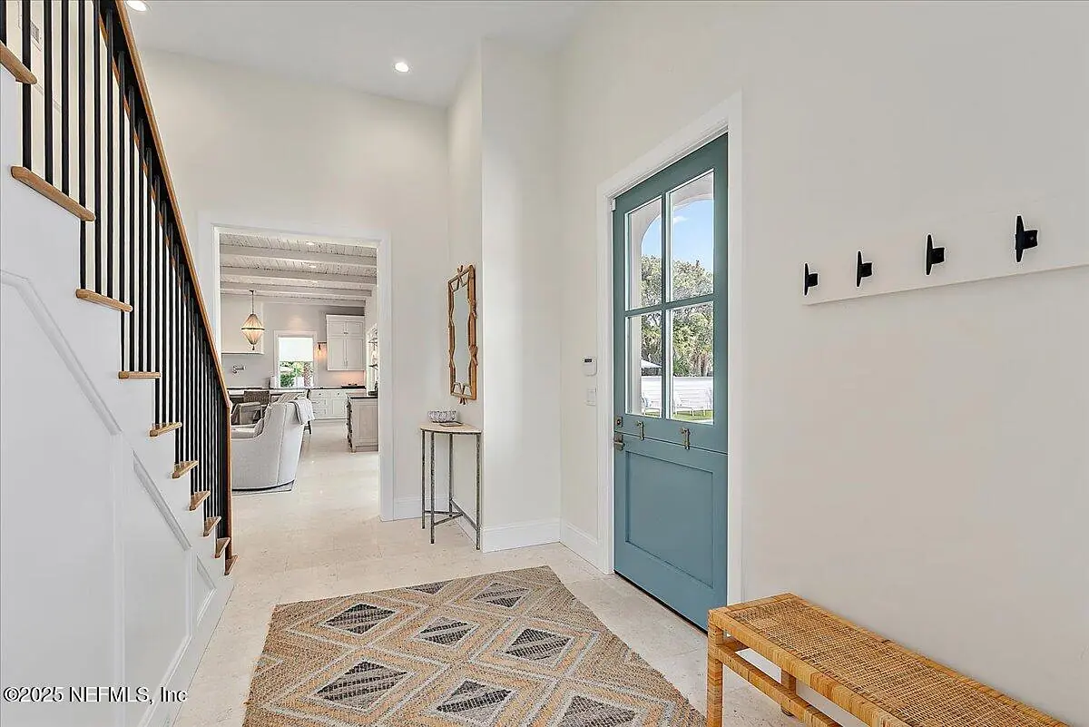 Foyer with blue door, woven bench, patterned rug, mirror, staircase, and wall hooks.
