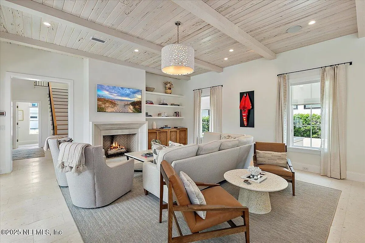 Living room with light-colored sofa, brown chairs, coffee table, fireplace, shelves, and large windows for light.