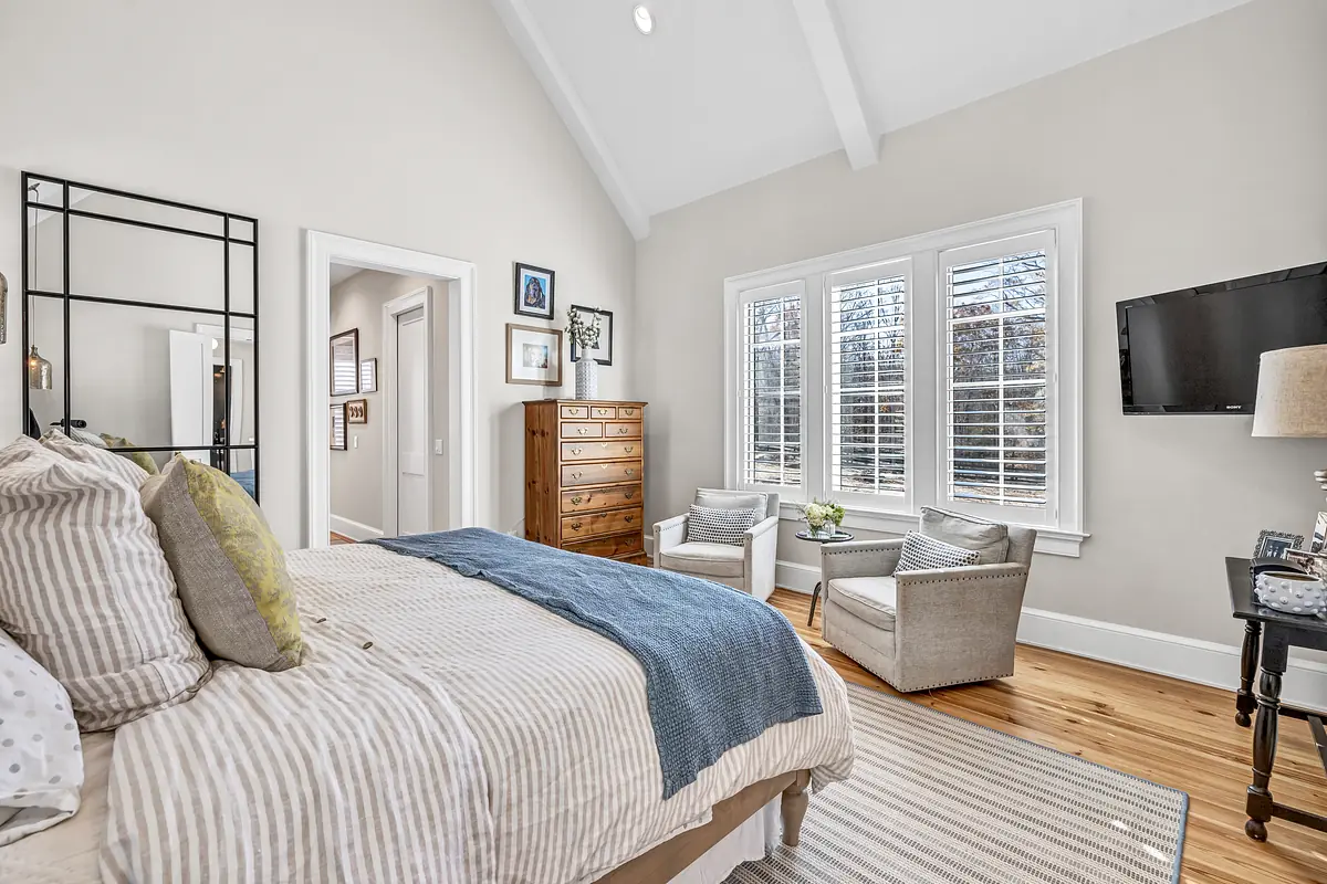 Bedroom with striped comforter, accent chairs, dresser, television, and vaulted ceilings.