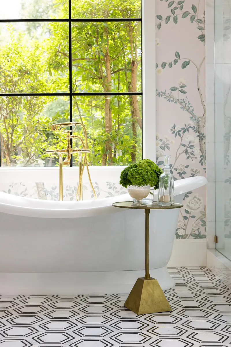 Bathroom with freestanding white bathtub, gold faucet, floral wallpaper, and geometric patterned tiles