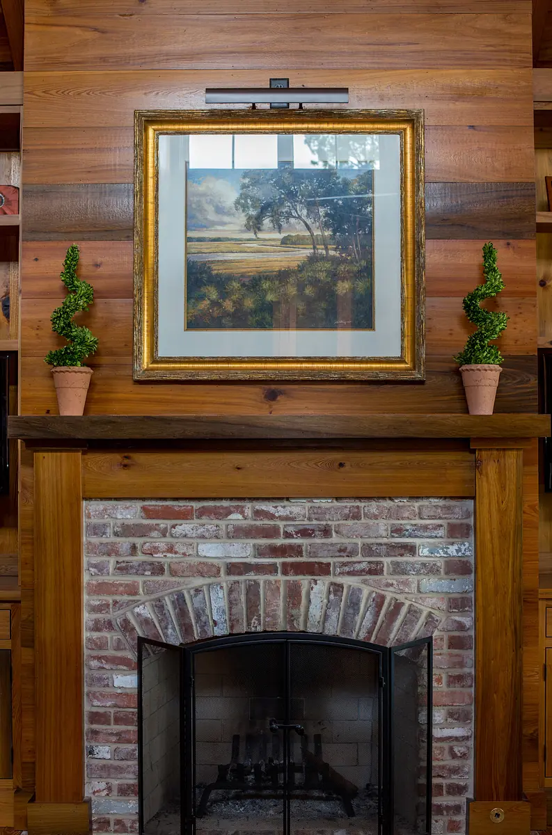 Living room with brick fireplace, wooden mantel, landscape painting, and potted topiary plants