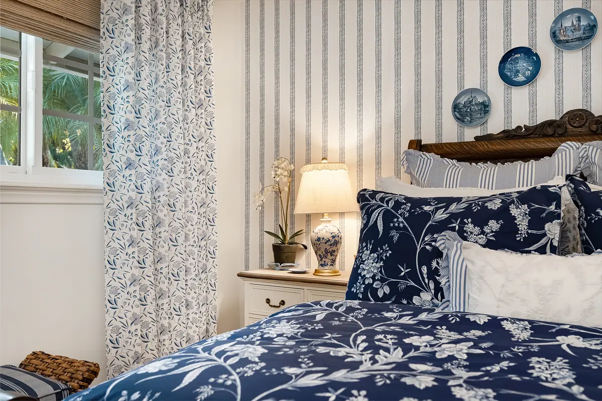 Bedroom with blue floral quilt, nightstands, lamp, striped wallpaper, and decorative plates.