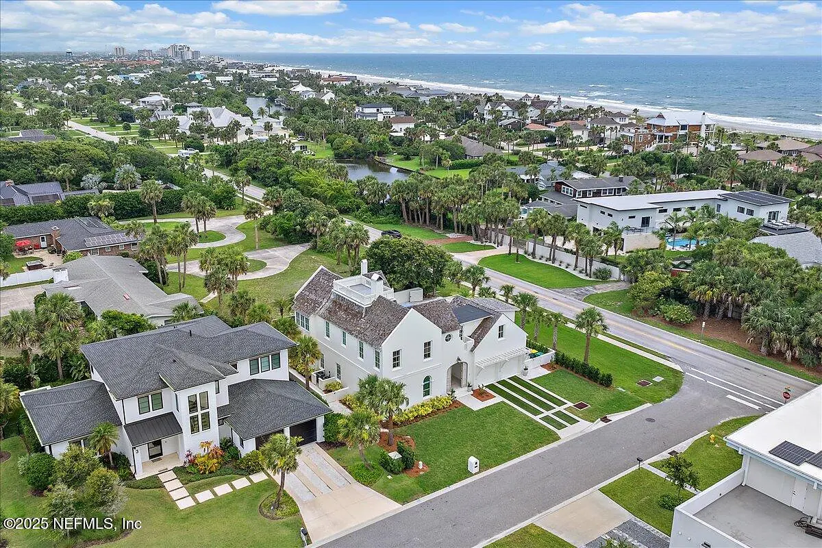 Exterior front view of two houses with green lawns, palm trees, and a street, overlooking the ocean.