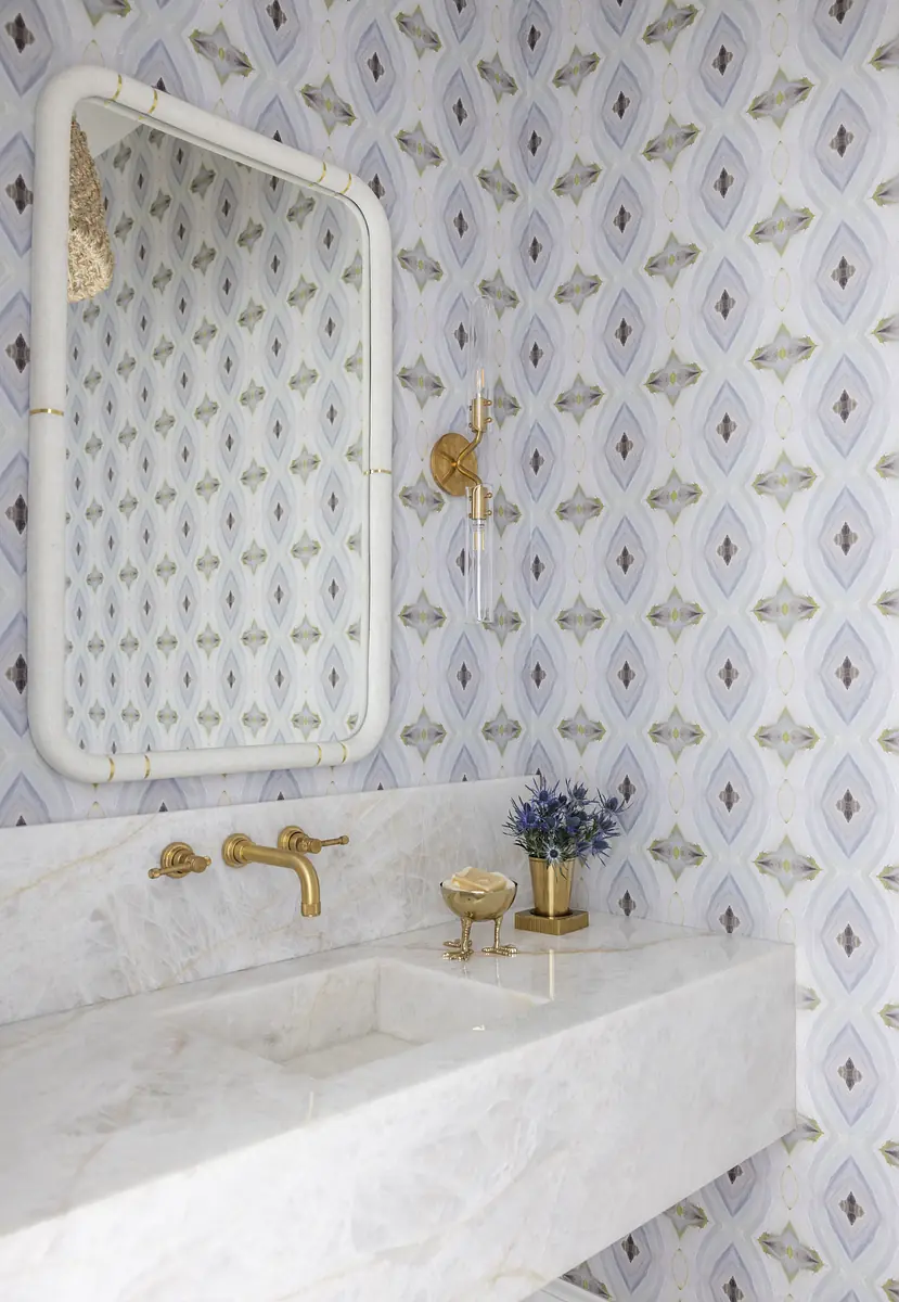 Bathroom with wall-mounted mirror, gold faucet, white marble sink, and patterned wallpaper with decorative accessories