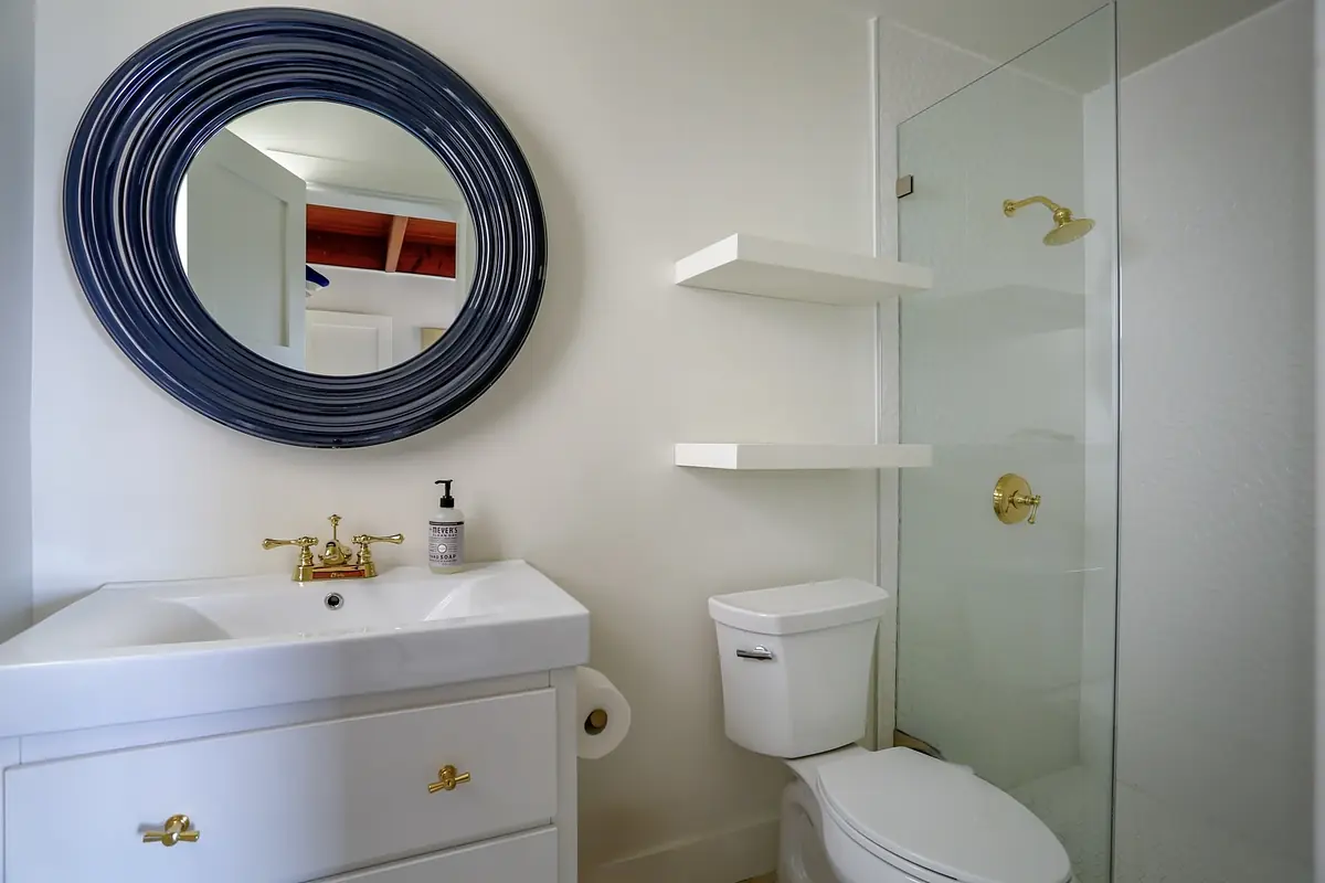 Bathroom with white vanity, gold faucet, round mirror, glass shower, and white toilet