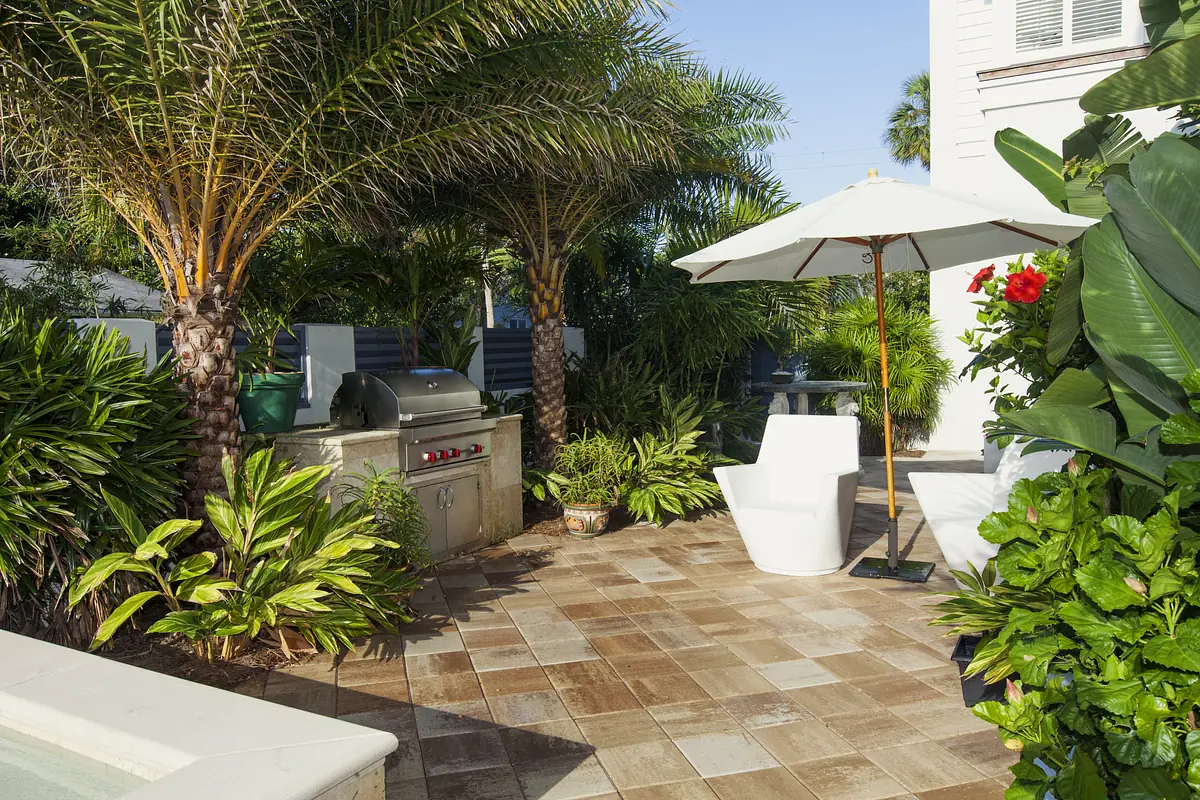 Patio with white chair, umbrella, stainless steel grill, and tropical plants on stone tile flooring.