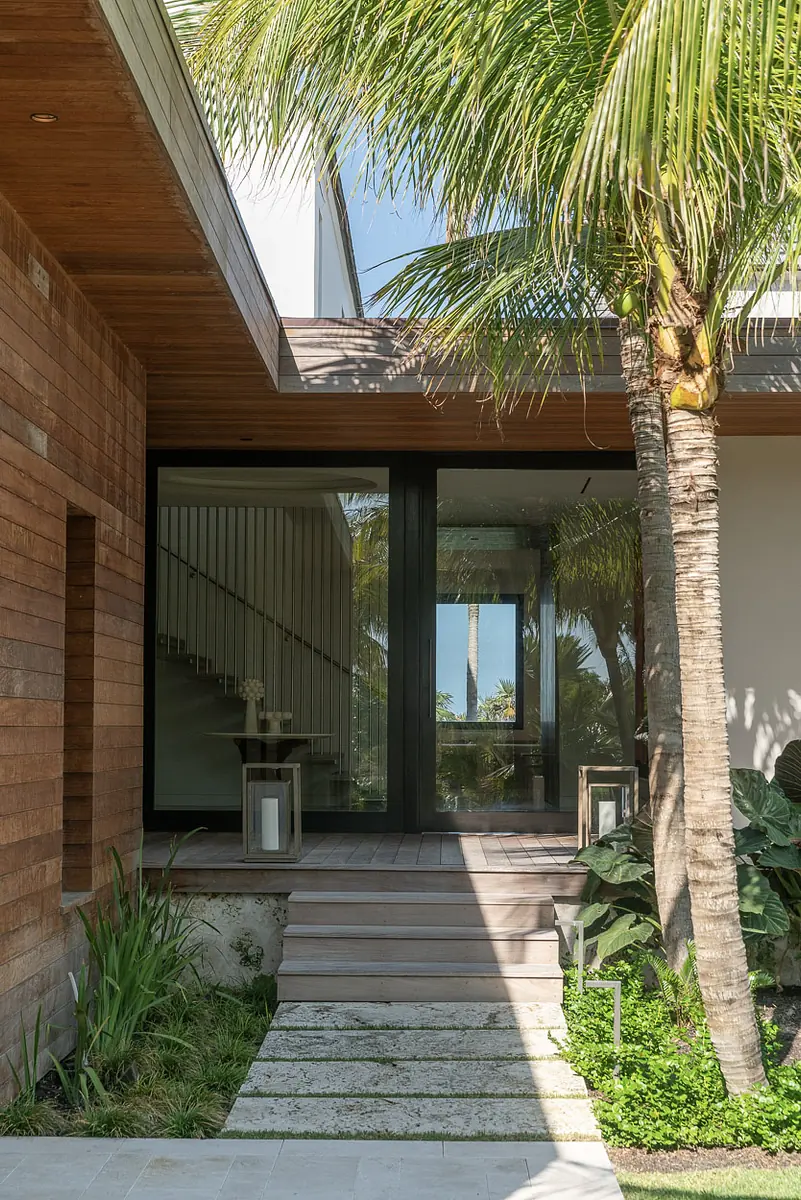 Exterior front entryway with wood-paneled wall, glass door, concrete pathway, and tropical plants
