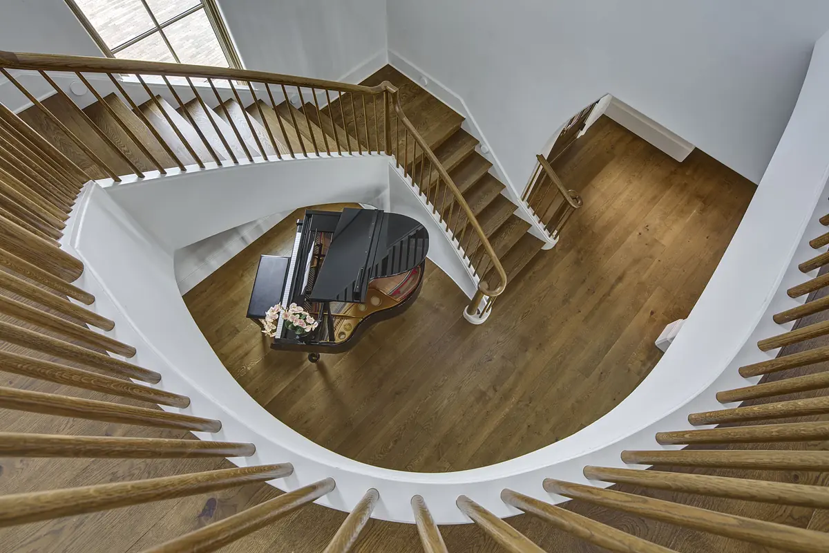 Curved staircase with grand piano on wooden floor and white walls illuminated by natural light