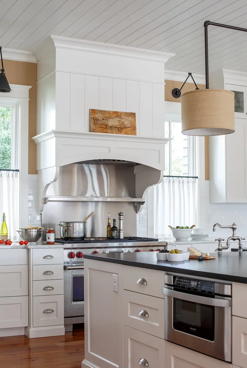Kitchen with white cabinetry, stainless steel range hood, black countertop, and large windows with fabric curtains.