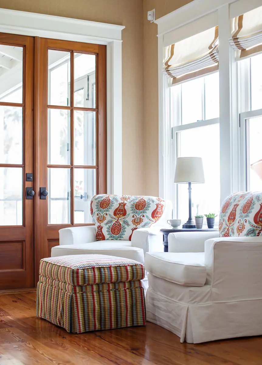 Living room with white chairs, striped ottoman, round table, lamp, and plants with natural light from large windows