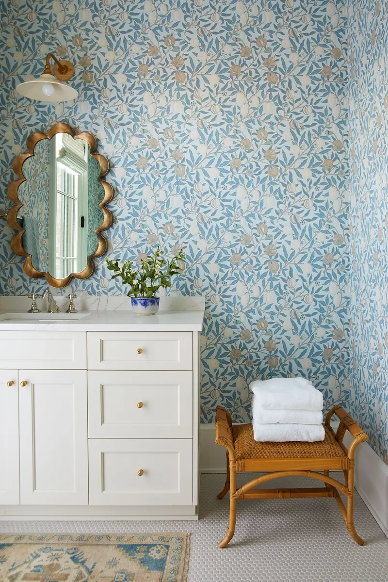 Bathroom with white vanity, round mirror, floral wallpaper, towels, and wicker stool.