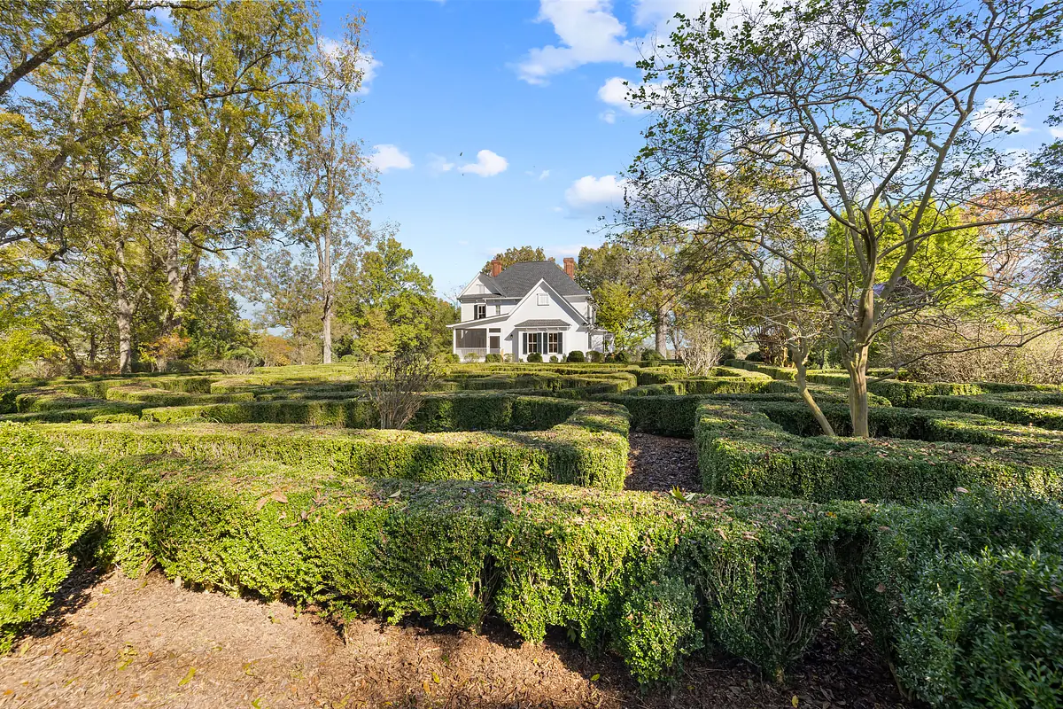 Garden with trimmed hedges forming a maze, trees, mulch, and view of house in background under partly cloudy sky.