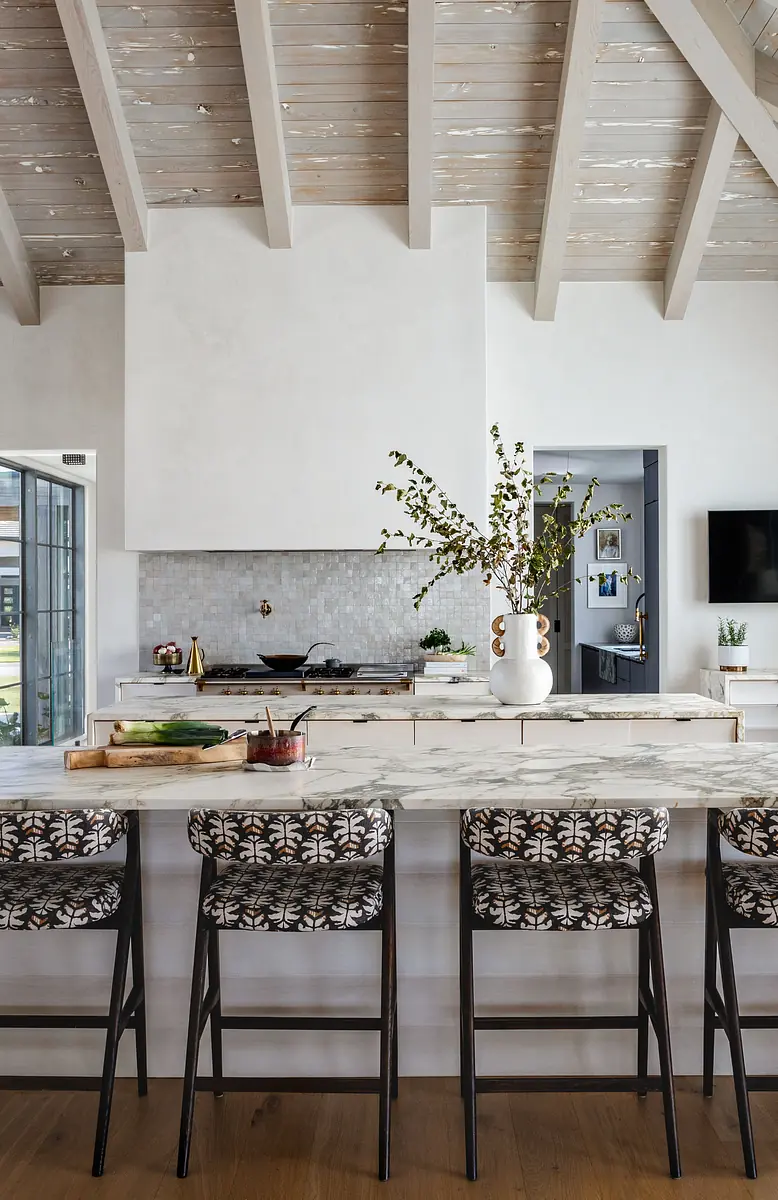 Kitchen with marble island, bar stools, backsplash, range hood, dark cabinetry, and wood beams on ceiling.