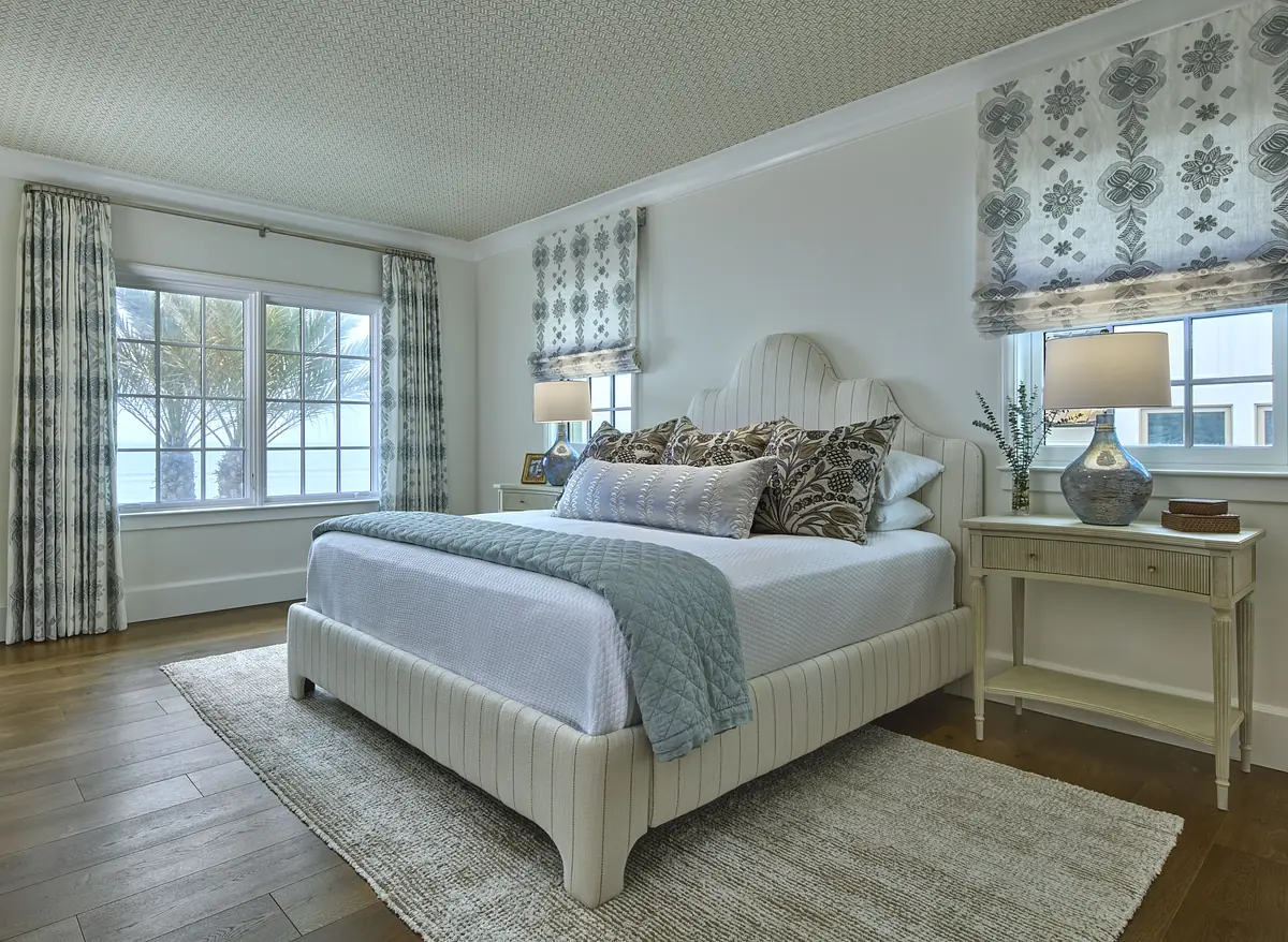 Bedroom with white bed, blue quilt, nightstands, lamps, and large windows with patterned curtains