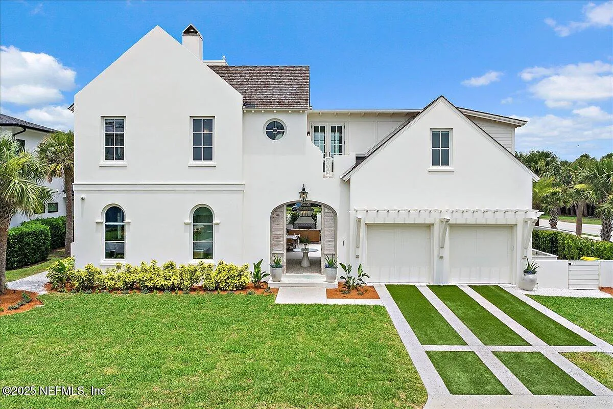 Exterior front view of a house with white stucco, arched windows, stone pathway, green lawn, and palm trees.