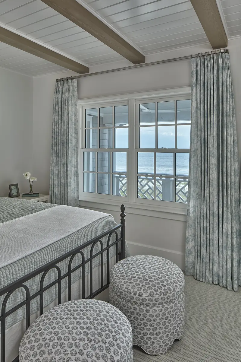 Bedroom with black metal bed frame, upholstered stools, sliding windows, and patterned drapes.