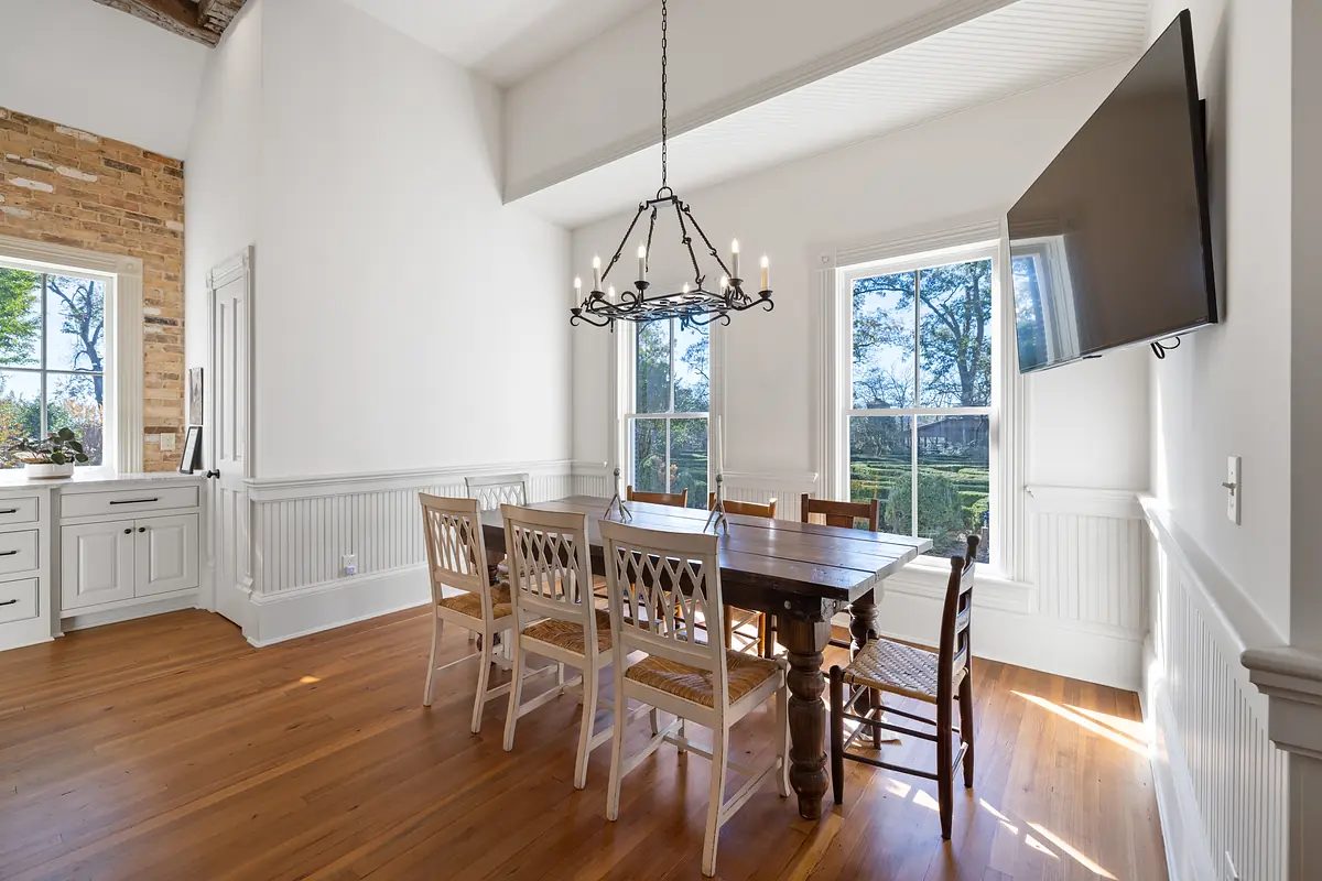 Dining room with wooden table, six chairs, chandelier, wainscoted walls, and large windows.