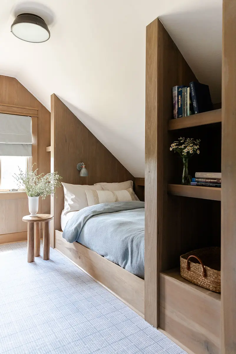 Bedroom with angled ceiling, gray blanket bed, wooden shelves, side table, and flower vase near a window