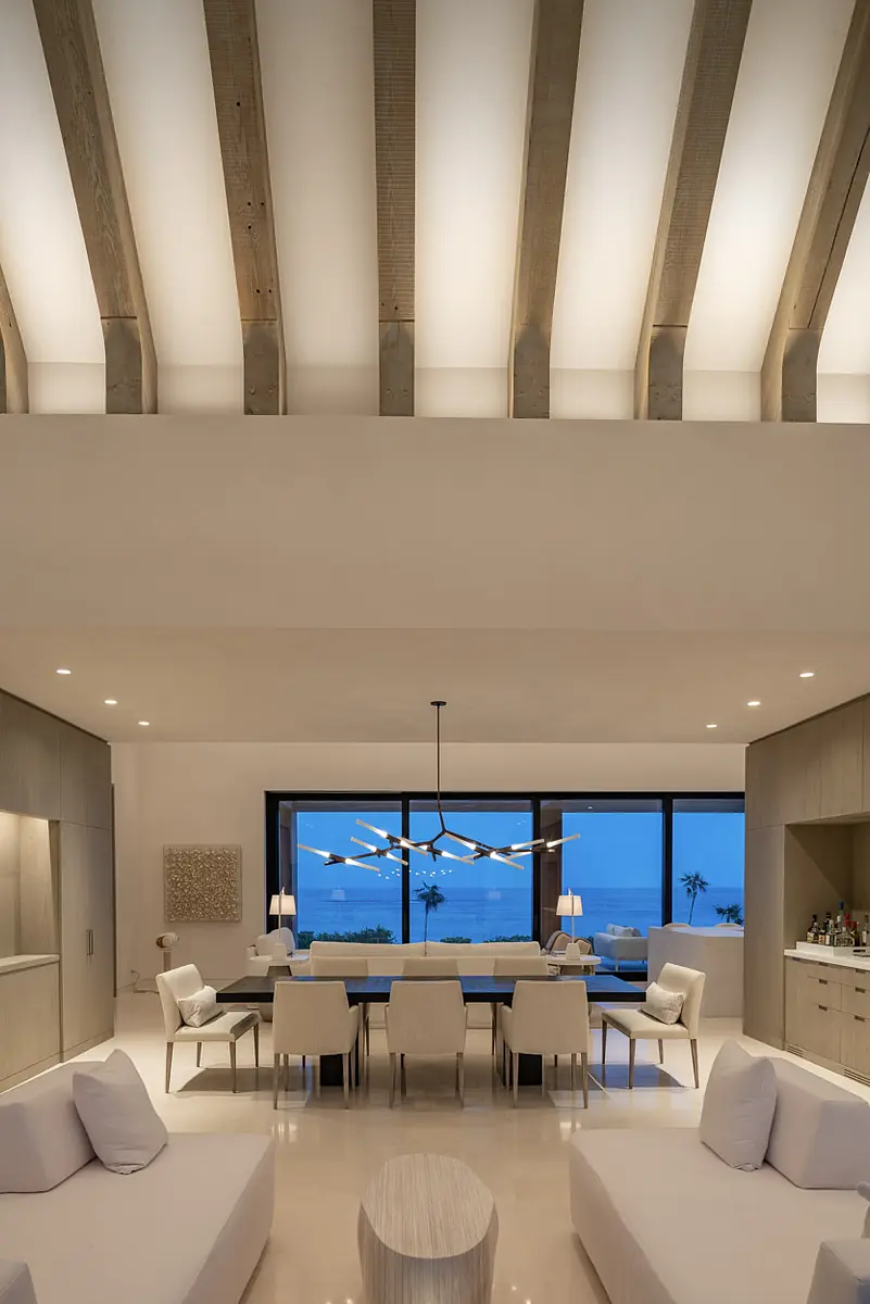 Living room with white sofas, circular table, dining table, white chairs, large window, and wooden beams ceiling.
