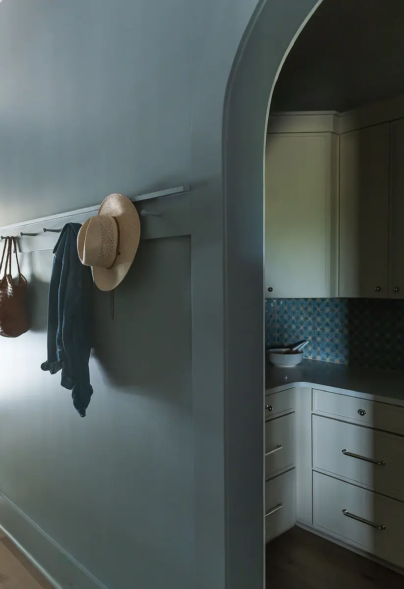 Mudroom with blue wall, coat rod, hats, and white cabinetry with blue tiled backsplash.