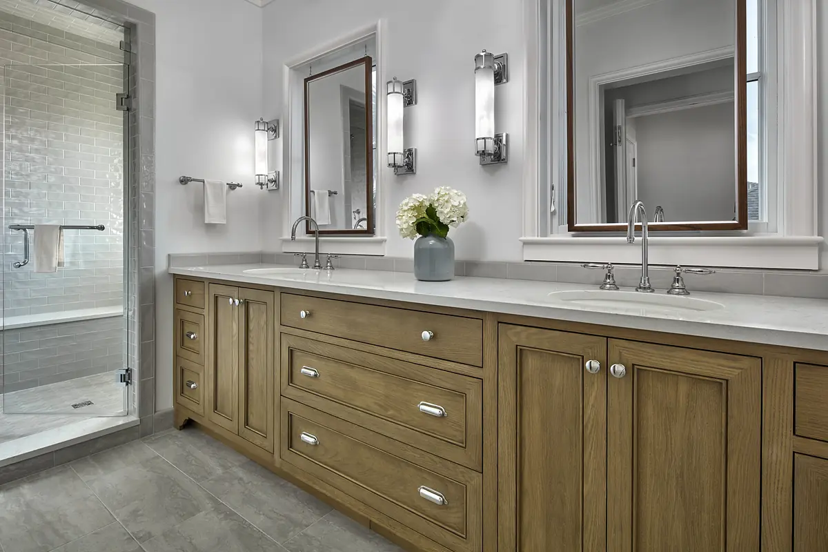 Bathroom with double sink vanity, wooden cabinetry, mirrors, wall sconces, and a glass shower enclosure.