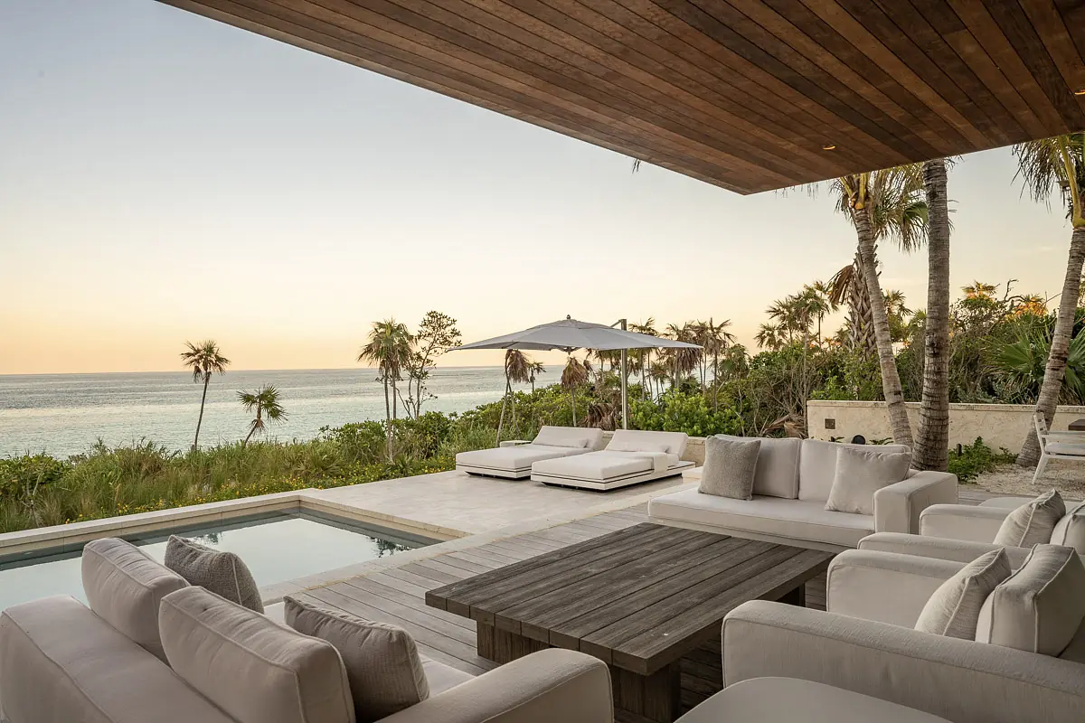 Patio with white outdoor sofas, wooden coffee table, umbrella, pool, and ocean view surrounded by tropical plants.