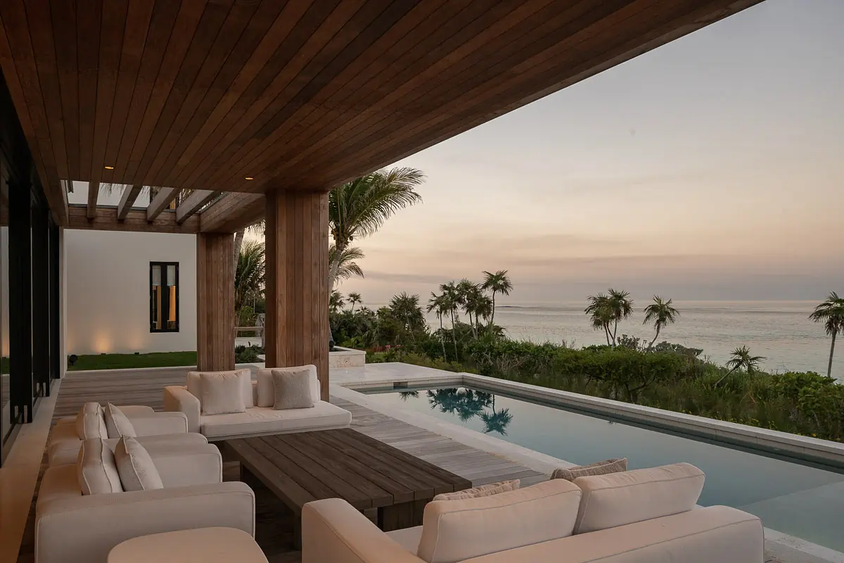 Deck with white sectional sofas, low wooden table, pool view, and landscaped garden with palm trees at sunset.