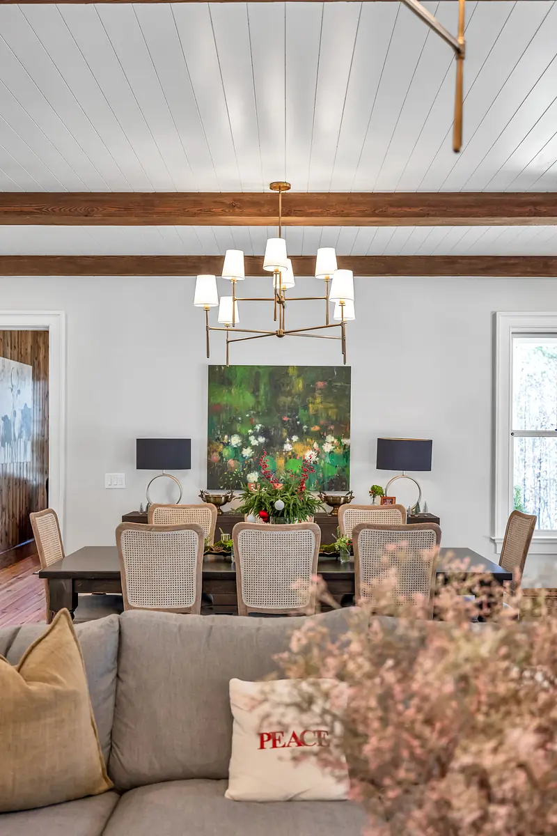 Dining room with large wooden table, floral painting, chairs, and beamed ceiling.