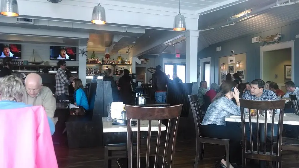 Dining room with tables, wooden chairs, patrons, televisions, nautical decor, and exposed beams.